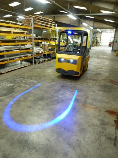 Tracteur électrique jaune circulant dans un entrepôt industriel, équipé de feux LED et d’un projecteur au sol en forme de U pour signaler sa trajectoire.