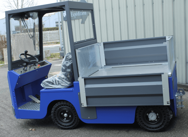 Tracteur électrique bleu à cabine ouverte, équipé d’un plateau de chargement arrière à ridelles métalliques, utilisé pour l’expédition de marchandises.